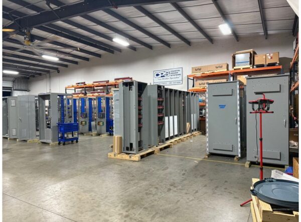 891Switchboard - Full warehouse CPSafe - Coastal Power Systems Industrial warehouse with rows of large gray electrical cabinets on pallets and blue rack shelving along the back wall, under a metal ceiling with bright lights. Sign reads PCS Coastal Systems.