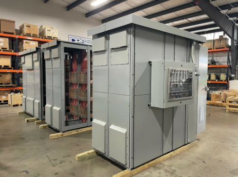 Two large gray industrial electrical enclosures on wooden pallets in a warehouse; the nearer unit features a control panel on its side.